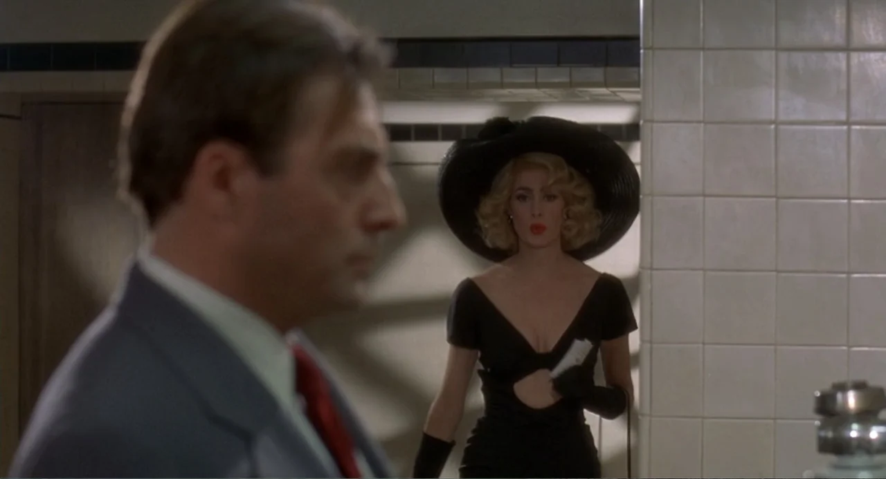 Fatal Instinct (1993) movie still: medium — A clean restroom with white tiled walls and a drop ceiling. A woman in a black dress stands in the…