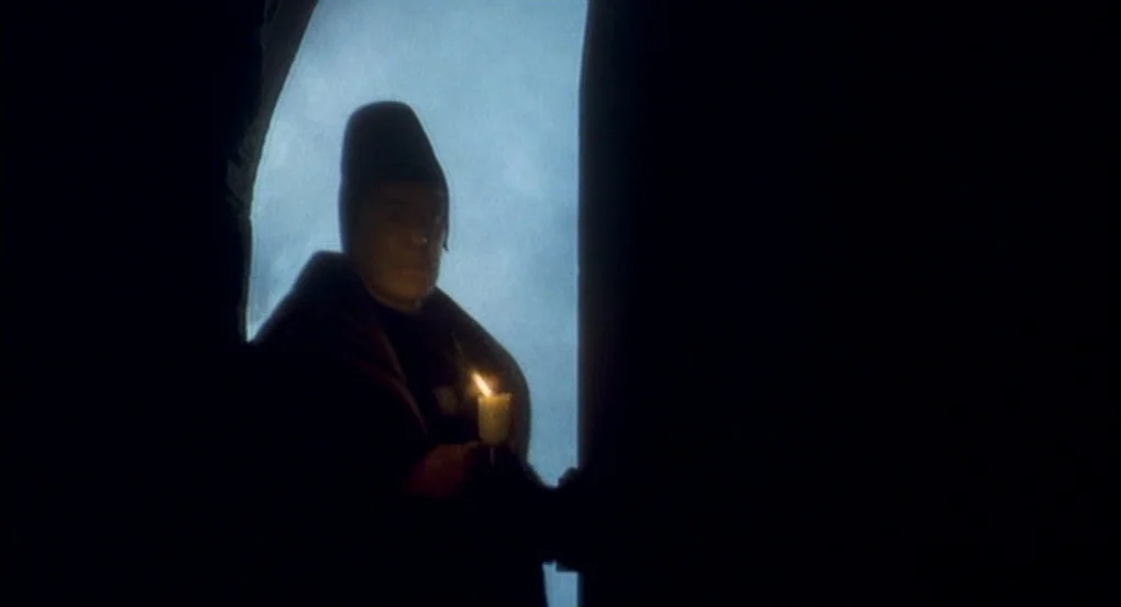 Jabberwocky (1977) movie still: medium — A dark stone archway framed against a dimly lit blue background. An elderly man in a dark cloak holds a…
