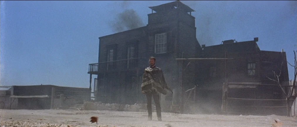 Fistful Of Dollars (1964) movie still: fullbody — A desolate western town features weathered wooden buildings under a clear, bright blue sky. A man…