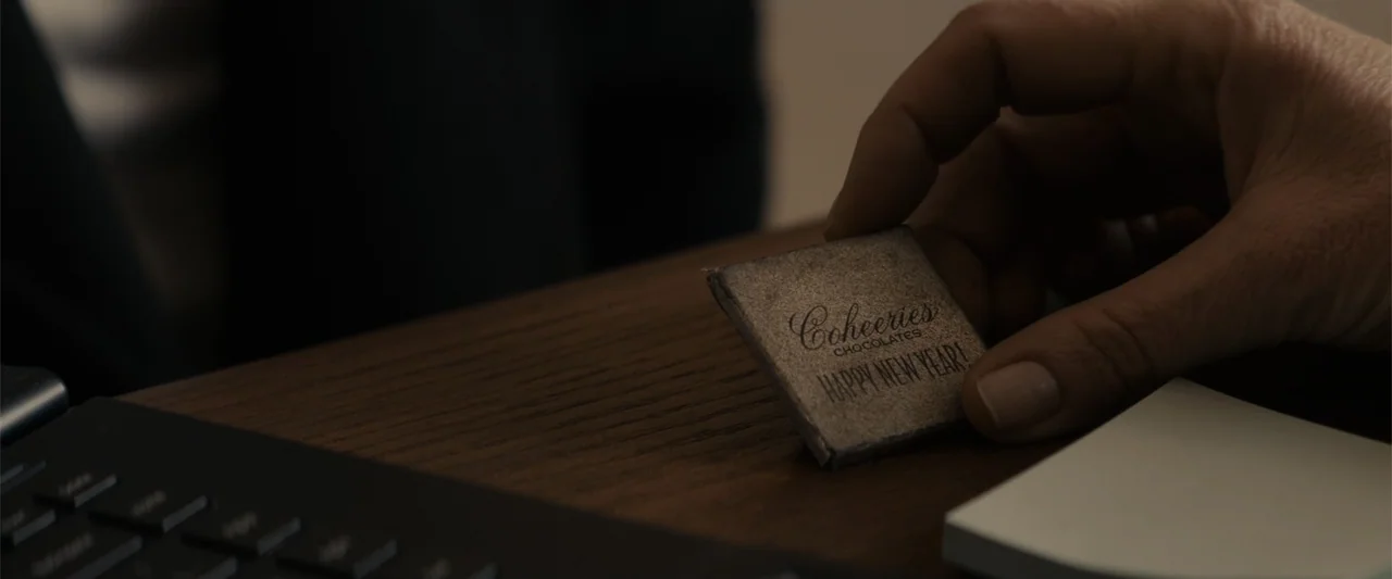 Winter's Tale (2014) movie still: closeup — A dark wooden desk holds a black keyboard and a small candy wrapper. A person's hand picks up the…
