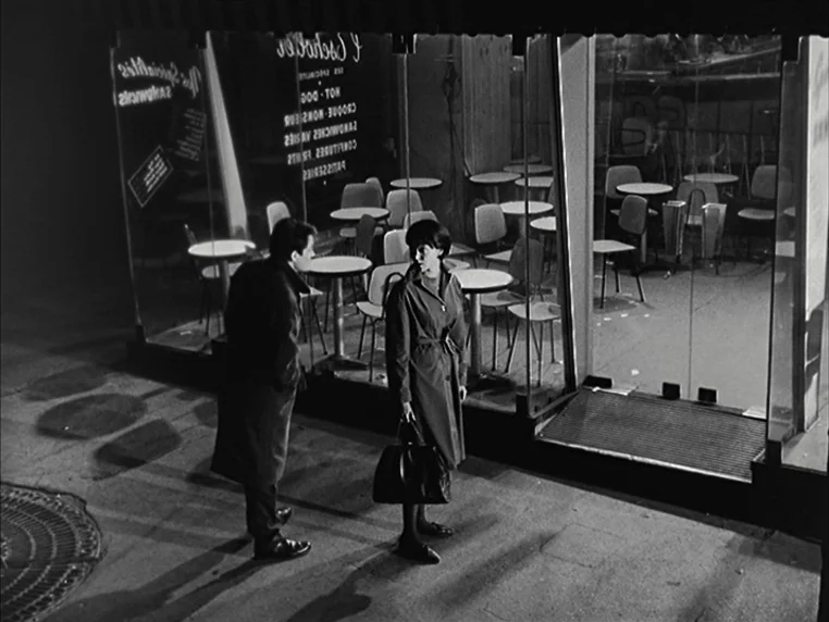 Vivre Sa Vie (1962) movie still: wide — A nighttime city sidewalk runs past the large glass facade of a quiet cafe. A man and a woman in trench…
