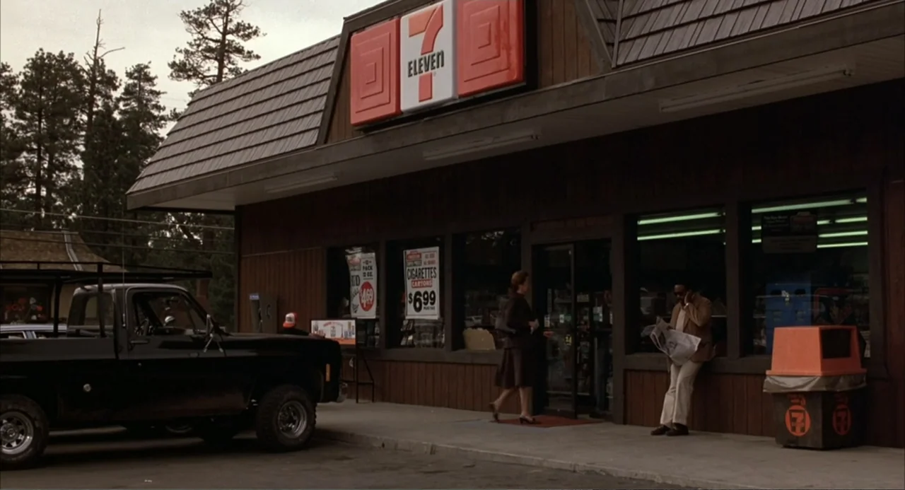 WarGames (1983) movie still: wide — A 1990s 7-Eleven store exterior features wood-paneled walls and a prominent orange sign. A woman in a brown coat…
