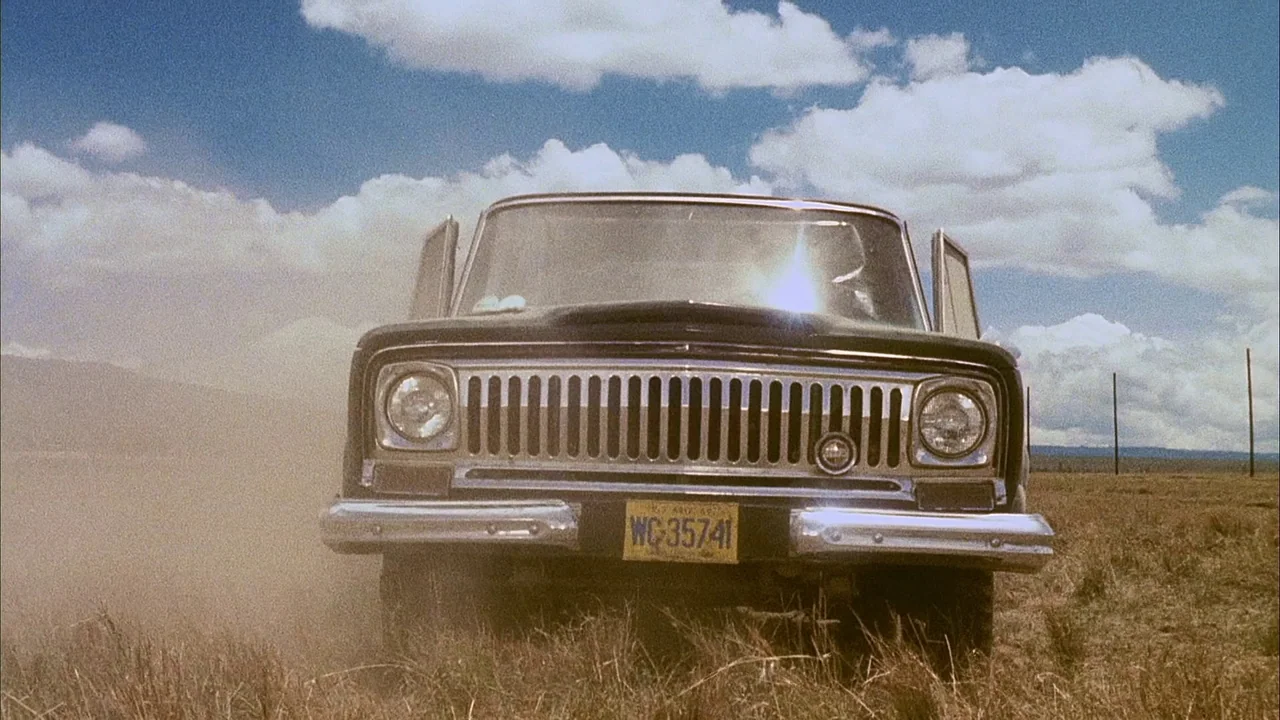 Phase IV (1974) movie still: medium — A dark-colored vintage SUV travels across a dry, grassy field under a bright sky. The vehicle is kicking up…