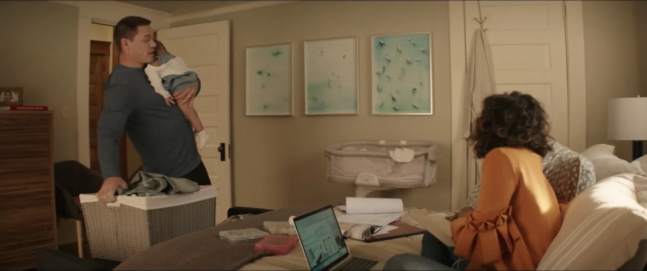 Blockers (2018) movie still: medium — A furnished nursery features a crib, framed artwork, and a bed with a laptop. A man holding a baby approaches…