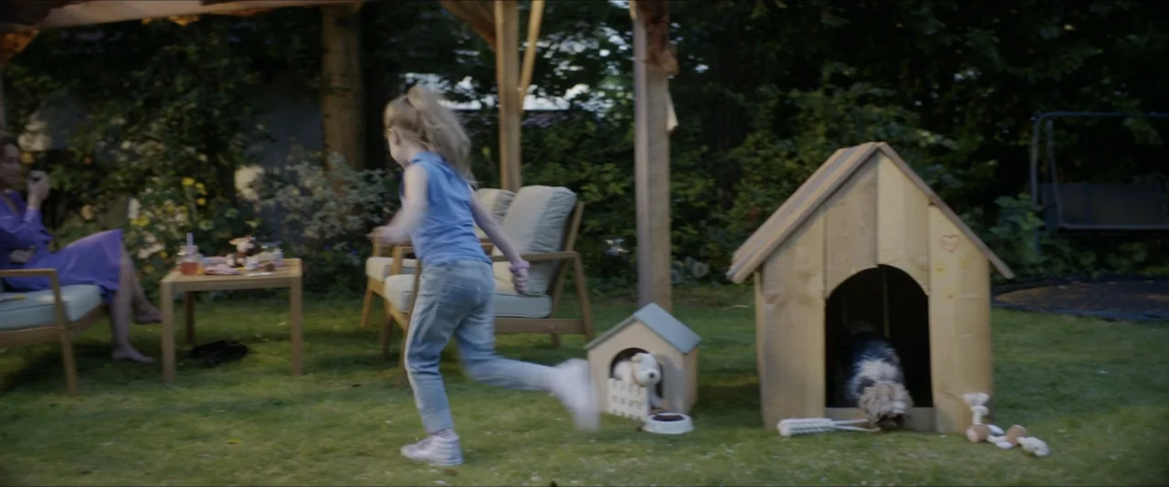 Black Lotus (2023) movie still: medium — A suburban backyard features a wooden dog house and patio furniture on green grass. A young girl in a blue…