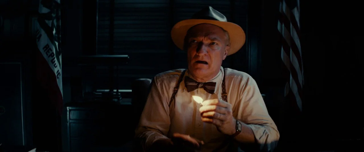 Gangster Squad (2013) movie still: medium — A dark, dimly lit 1940s office featuring a California state flag and an American flag. A man in a hat…