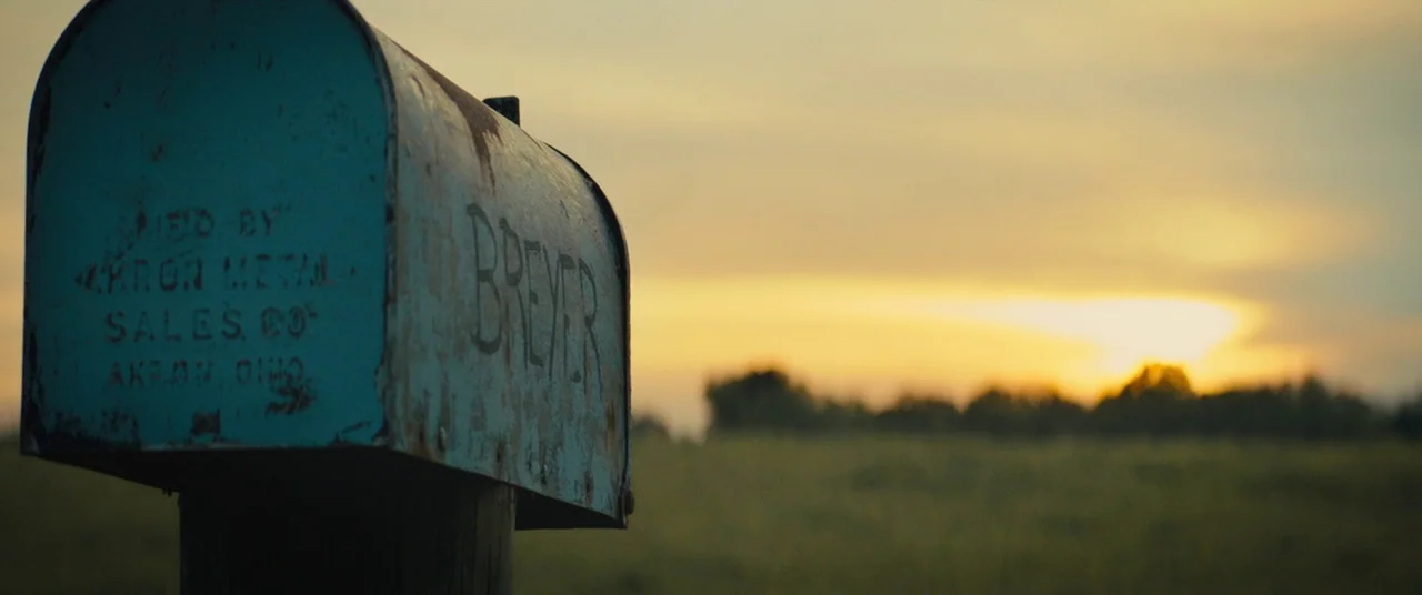 Brightburn (2019) movie still: closeup — A weathered, blue metal mailbox stands in the foreground against a rural landscape. The sun sets behind a…
