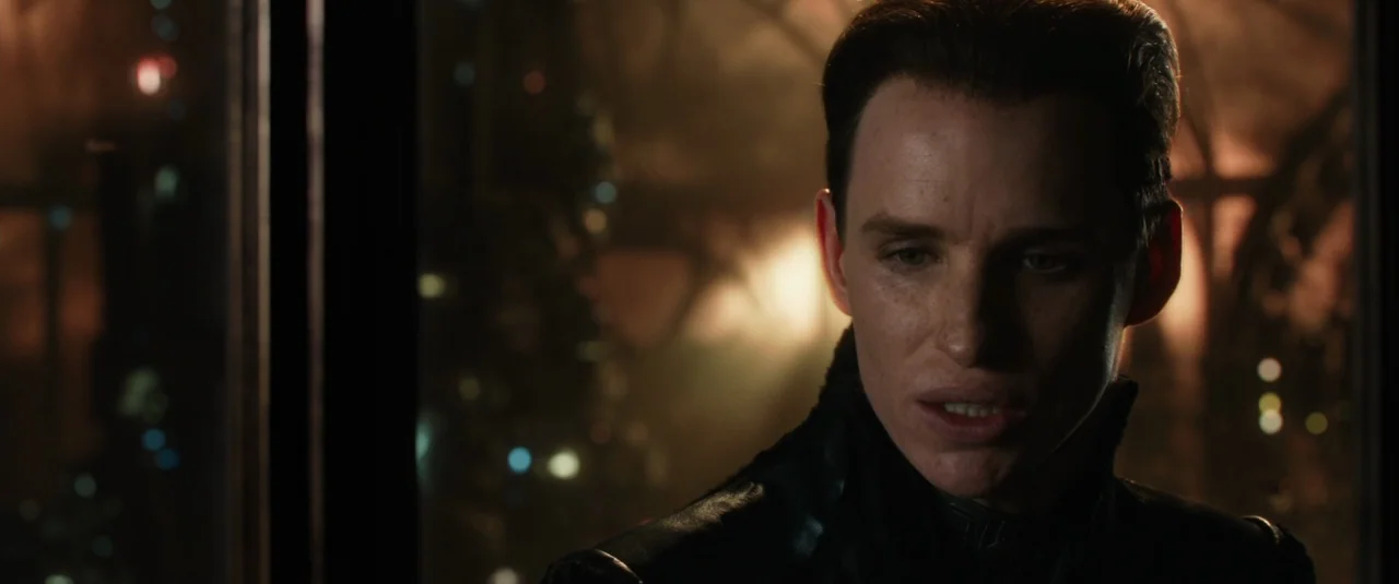 Jupiter Ascending (2015) movie still: closeup — A dimly lit room overlooks a hazy, indistinct cityscape at night through glass. A man in a…