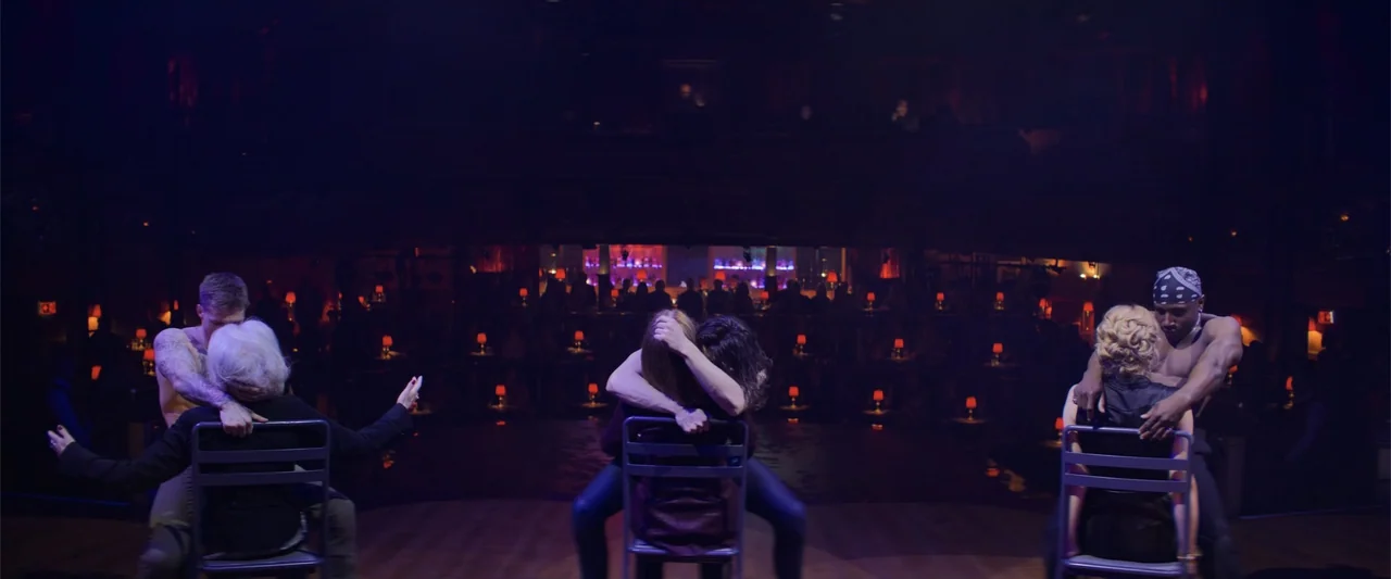 Magic Mike's Last Dance (2023) movie still: wide — A dimly lit nightclub stage contains three couples sitting on individual metal chairs in…