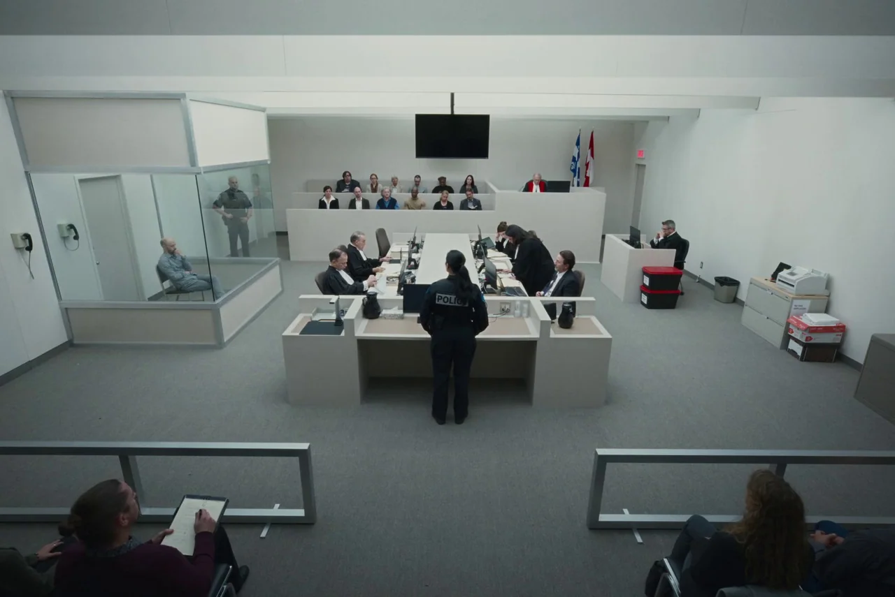 Red Rooms (2023) movie still: wide — A modern, sterile courtroom features white walls and a large central glass prisoner enclosure. People in legal…