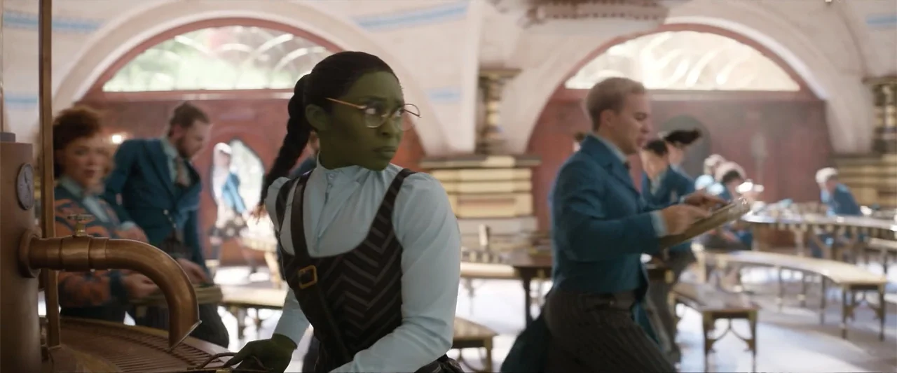 Wicked (2024) movie still: medium — An ornate interior dining hall features high arched windows and long wooden benches. A green-skinned woman…