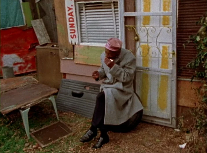Desperate Living (1977) movie still: fullbody — A cluttered backyard features a makeshift seating area composed of a car tire. An elderly man in a…