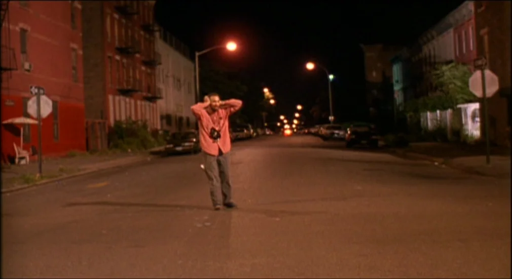 Do The Right Thing (1989) movie still: wide — An empty residential street at night is lined with brick buildings and tall trees. A lone man stands…