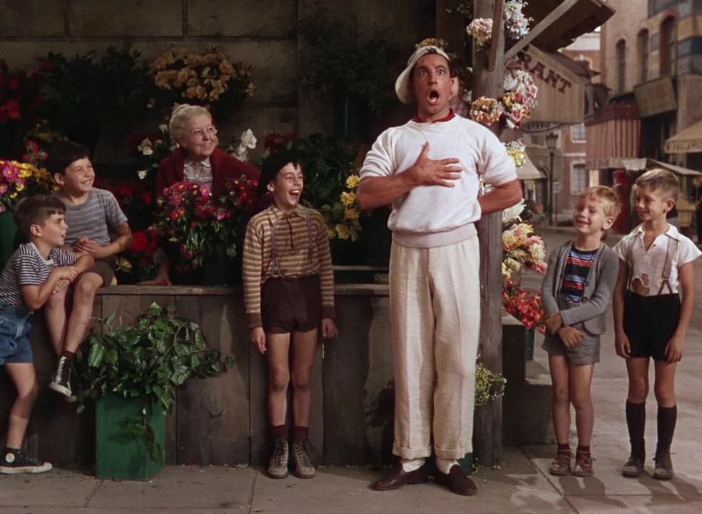 An American In Paris (1951) movie still: fullbody — A sidewalk flower stall displays various colorful blooms in containers against a wooden counter.…