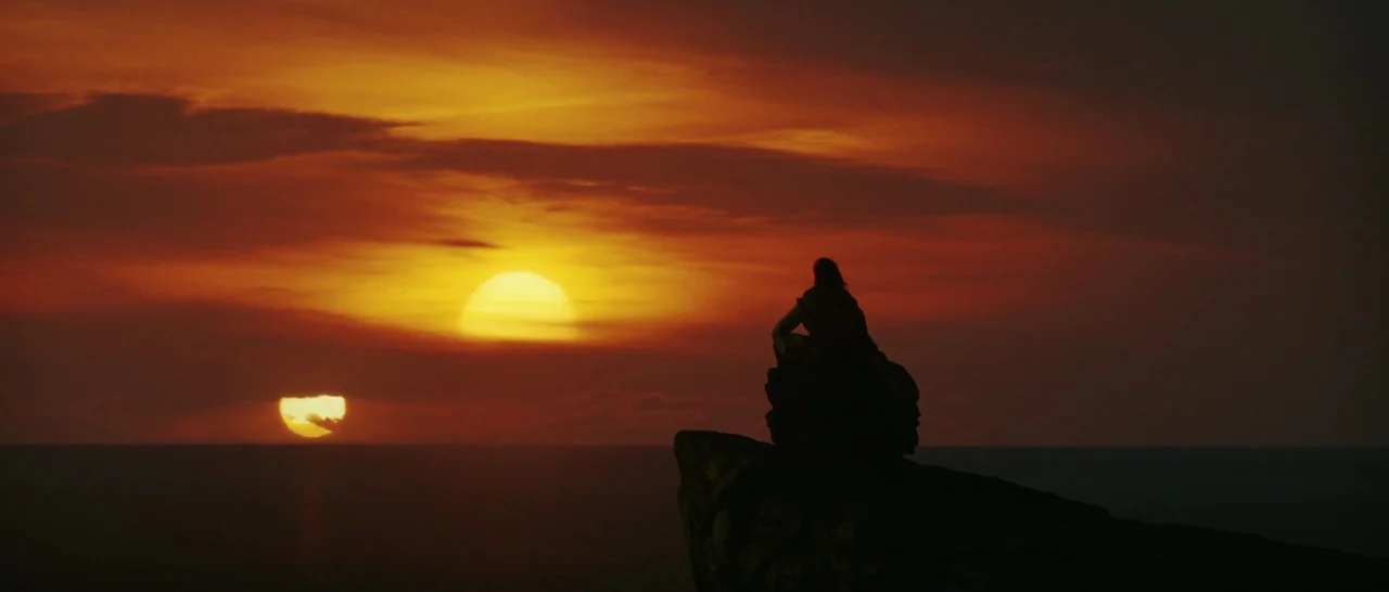 Star Wars: The Last Jedi (2017) movie still: wide — A dark rocky cliff juts into a vast horizon under a deep orange sky. A man sits motionless on…