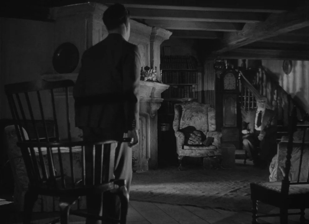 Dead of Night (1945) movie still: medium — A dimly lit traditional room features heavy furniture, a fireplace, and a grandfather clock. A man stands…