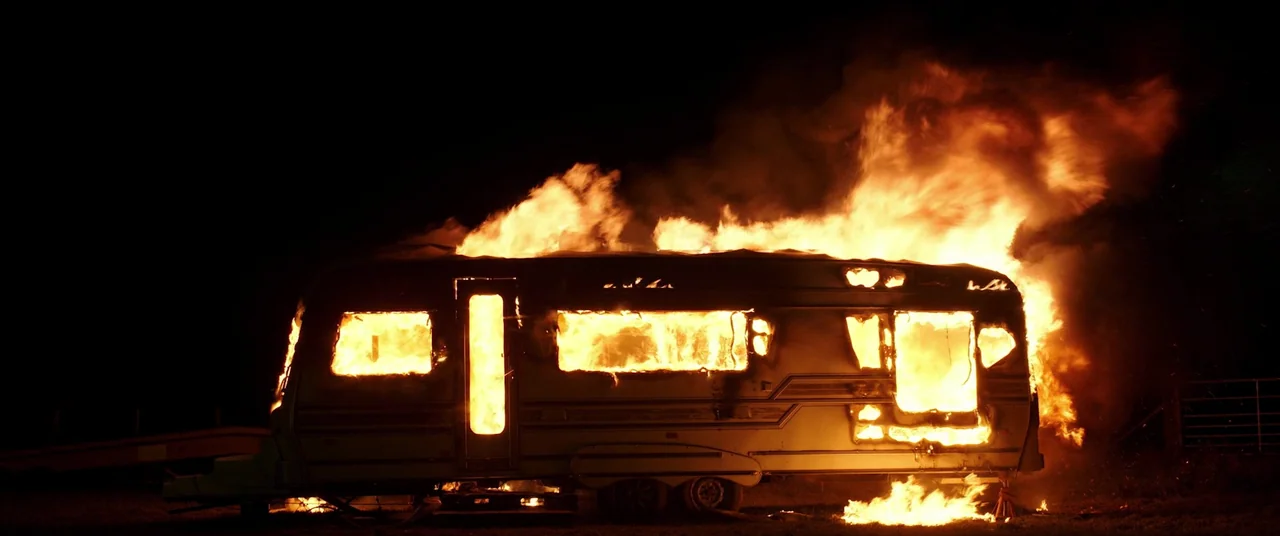 Bull (2021) movie still: wide — A travel trailer is engulfed in flames in an open dark field at night. Thick fire consumes the roof and windows of…