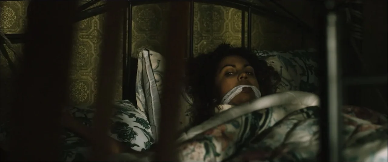 I'll Take Your Dead (2018) movie still: closeup — A dimly lit room features a woman lying in bed against floral wallpapered walls. The woman is…