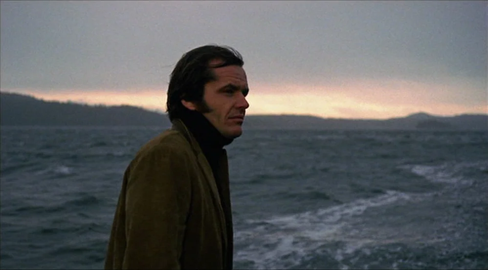 Five Easy Pieces (1970) movie still: medium — A man stands on a windy, overcast beach with a grey ocean in the distance. He gazes off-camera toward…