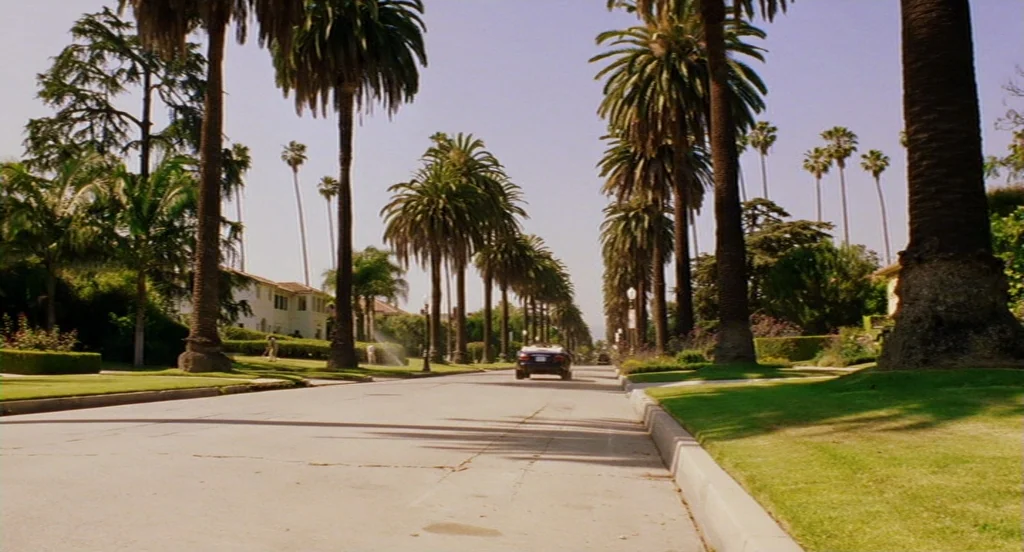 Intolerable Cruelty (2003) movie still: wide — A residential street is lined with tall palm trees and large suburban houses. A dark convertible car…
