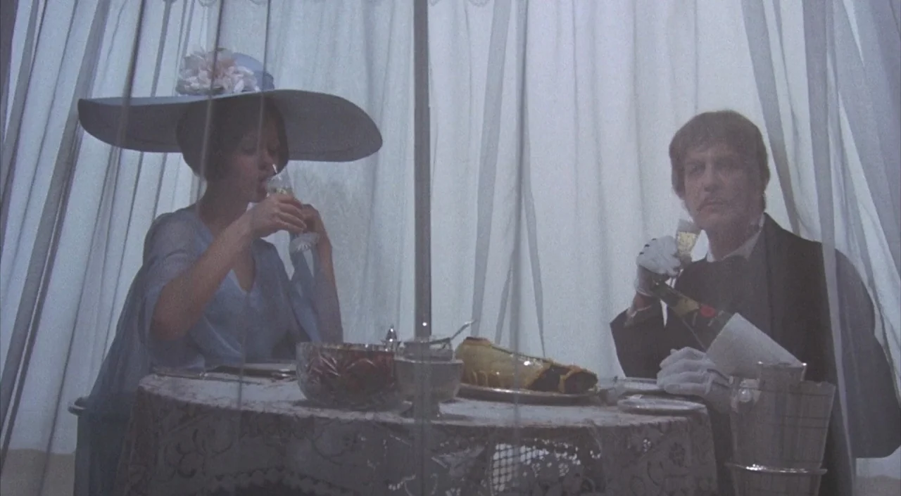 Dr. Phibes Rises Again (1972) movie still: medium — A lace-covered table is set with crystal and wine behind a layer of curtains. A woman in a wide…