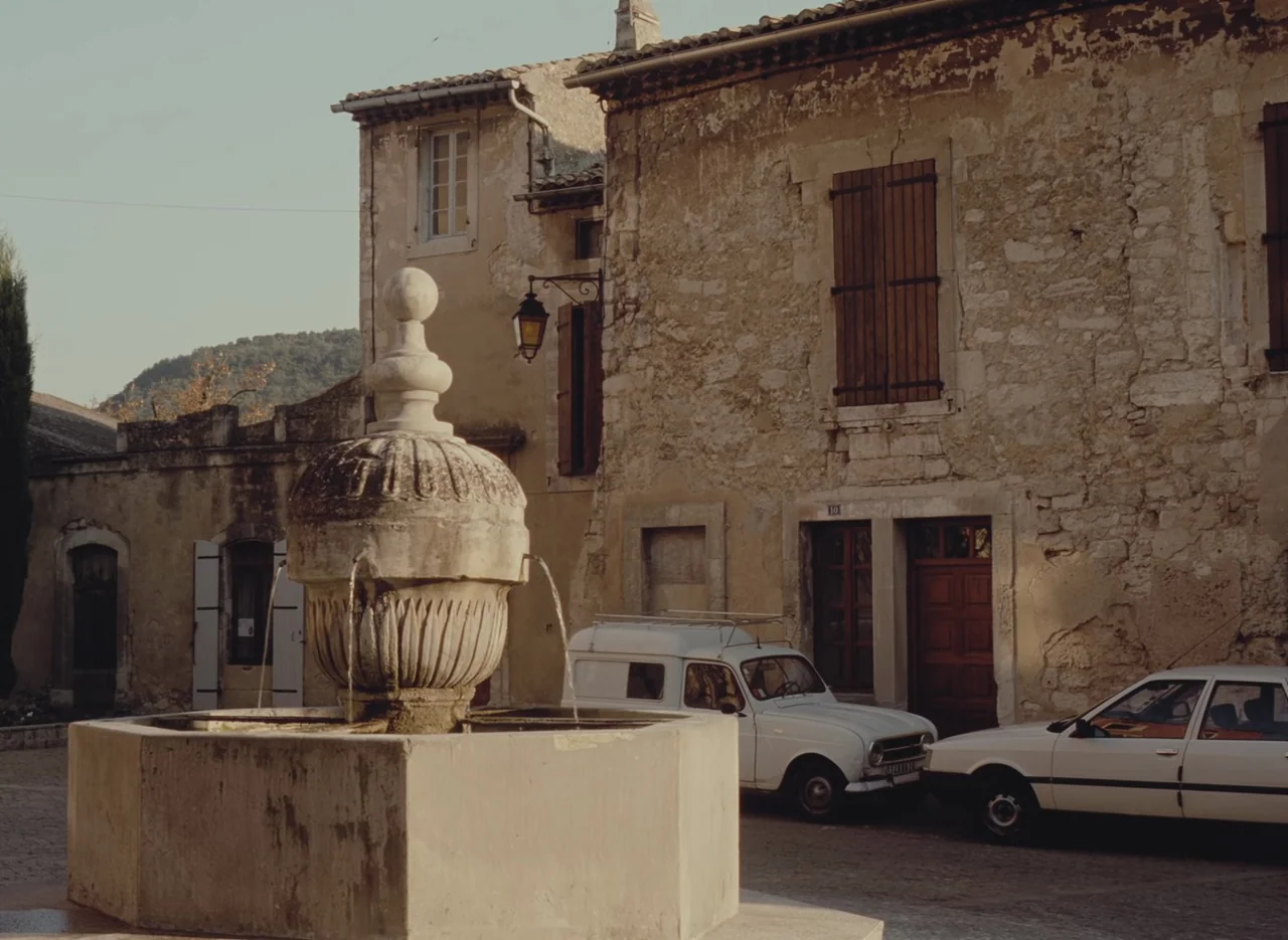 Autumn Tale (1998) movie still: wide — A stone square in a historic French village features an ornate fountain basin. Two vintage white cars are…