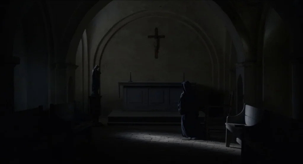 The Innocents (2016) movie still: wide — A dark stone church interior features stone arches, a central altar, and a crucifix. A single hooded figure…