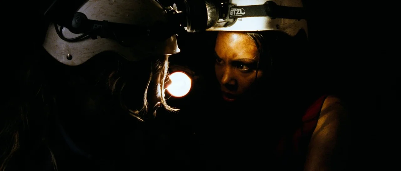 The Descent (2005) movie still: medium — Two individuals wearing caving helmets and headlamps look at each other in darkness. A bright beam from a…