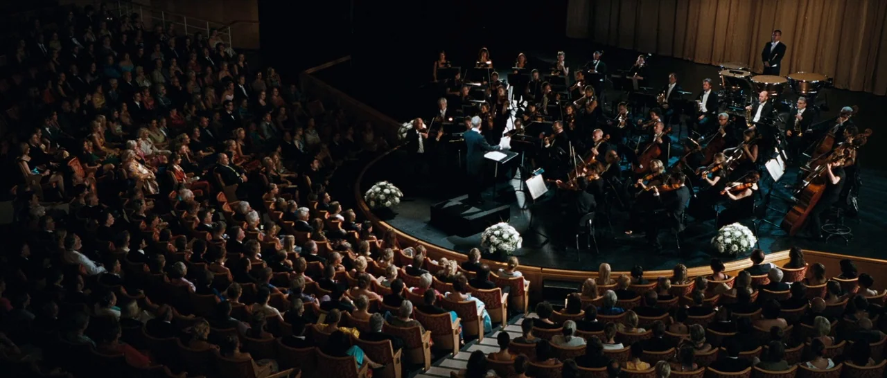 Sound of Noise (2010) movie still: wide — A crowded concert hall viewed from a high balcony vantage point. A full orchestra plays on a lit stage…