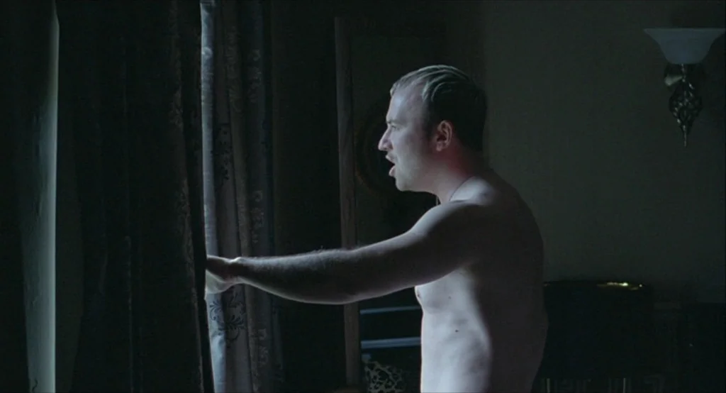 Looking for Eric (2009) movie still: medium — A dark, dimly lit space with a window partially covered by heavy drapes. A shirtless man stands by the…