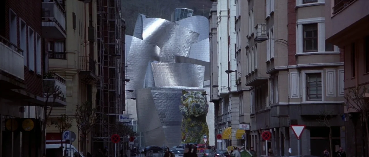 The World is Not Enough (1999) movie still: establishing — A narrow urban street leads toward a large, shimmering titanium-clad contemporary…