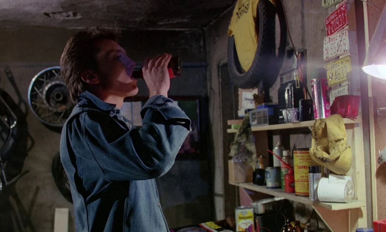 Rabid (1977) movie still: medium — A cluttered garage contains shelves filled with automotive supplies, tires, and old license plates. A man wearing…