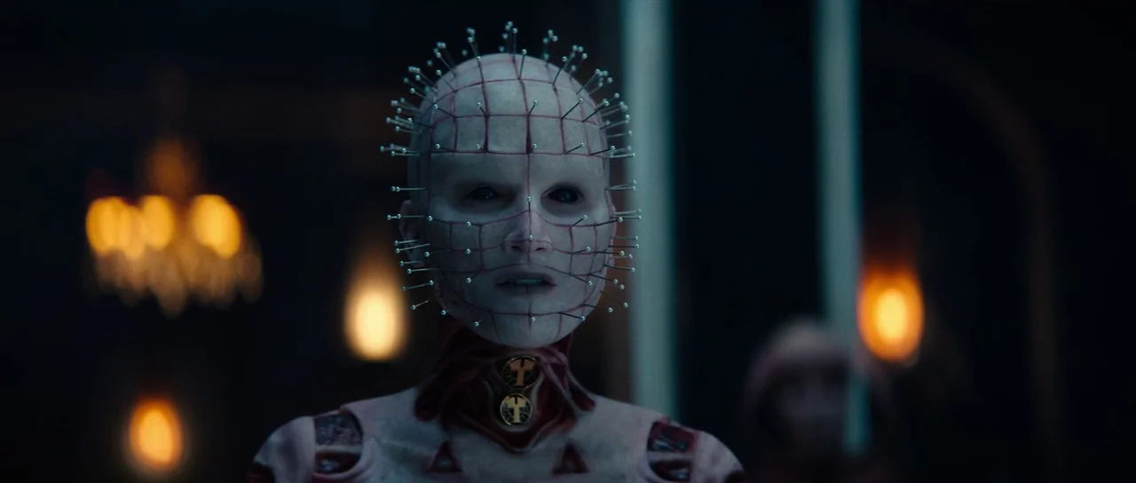 Hellraiser (2022) movie still: medium — A dimly lit chamber features blurred golden light sources in the out-of-focus background. A character with a…