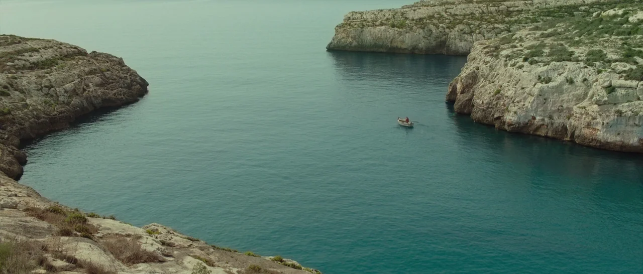 By The Sea (2015) movie still: wide — A secluded coastal inlet is enclosed by jagged, light-colored limestone cliffs and vegetation. A lone…