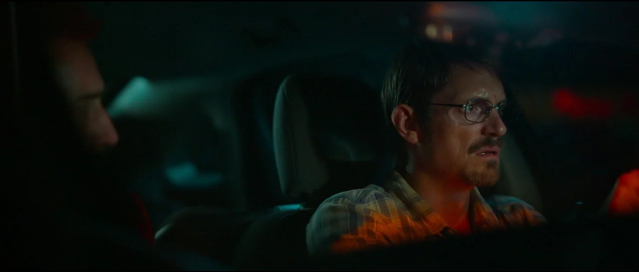 Sympathy For The Devil (2023) movie still: medium — The interior of a moving vehicle at night with dark surroundings. A man with glasses sits in the…