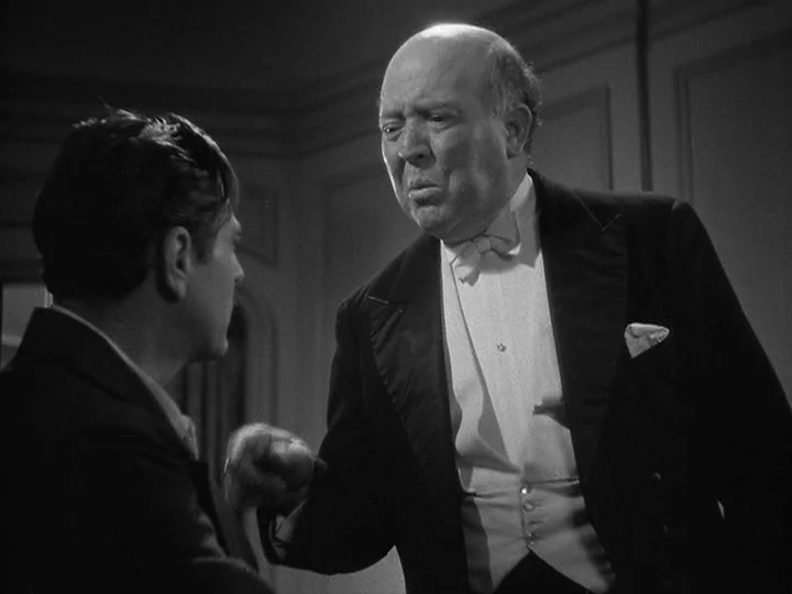 42nd Street (1933) movie still: medium — A dimly lit room with wood-paneled walls features two men in formal evening attire. The older man stands…