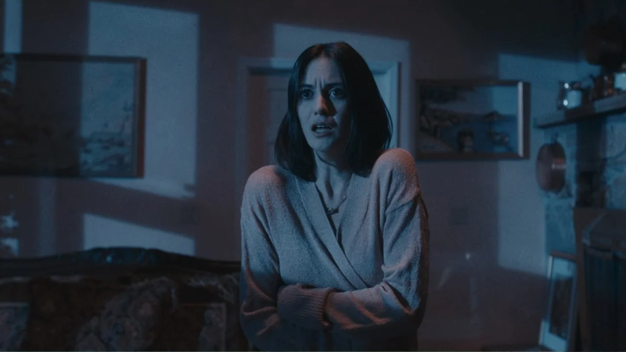Terrifier 2 (2022) movie still: medium — A dimly lit room with framed artwork on the walls and a fireplace. A woman in a knit sweater stands in the…