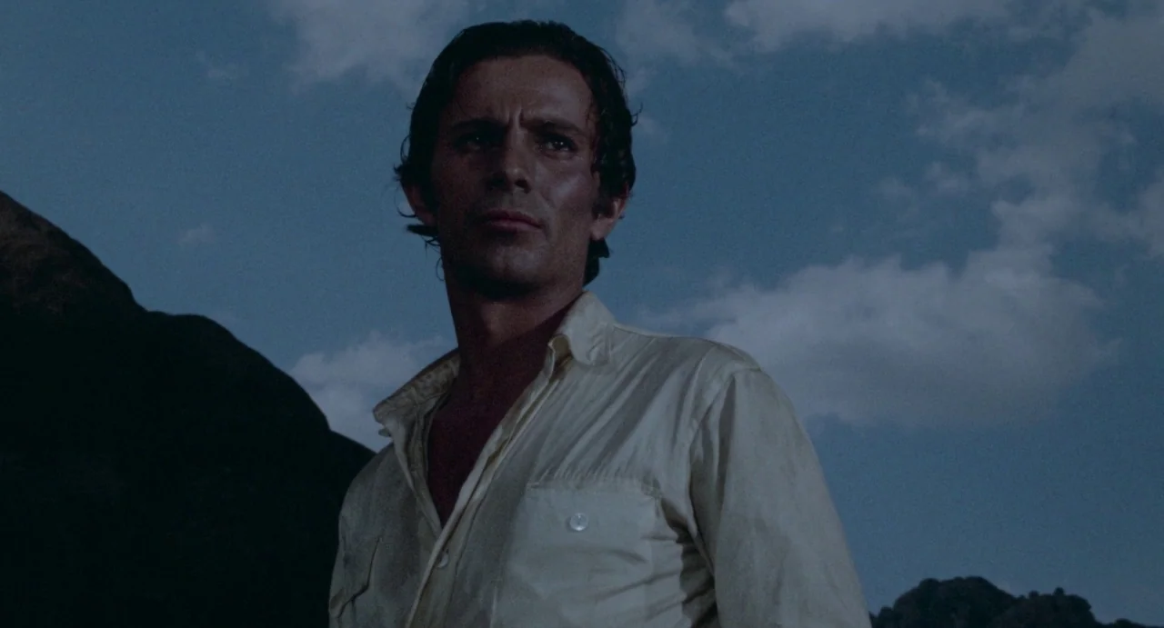 The Hellbenders (1967) movie still: medium — A man stands outdoors against a twilight sky with darkened hills in the background. He stands in…