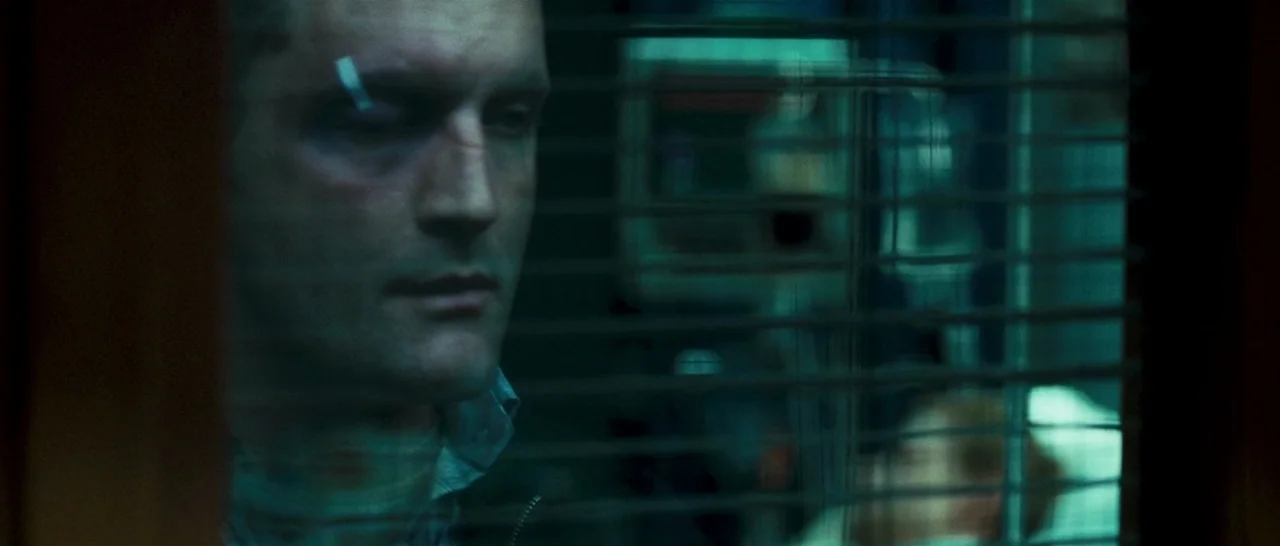 Green Street (2005) movie still: closeup — A dimly lit hospital hallway viewed through a glass panel with horizontal blinds. A man stands behind the…