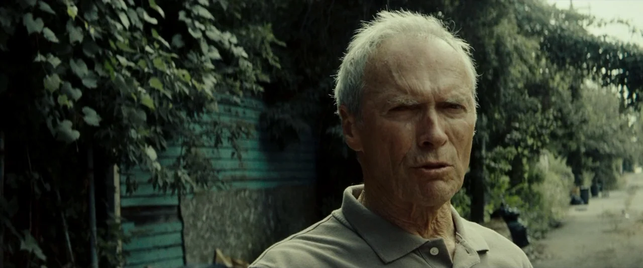 Gran Torino (2008) movie still: medium — A residential street lined with overgrown bushes and a turquoise wooden fence. An elderly man in a polo…