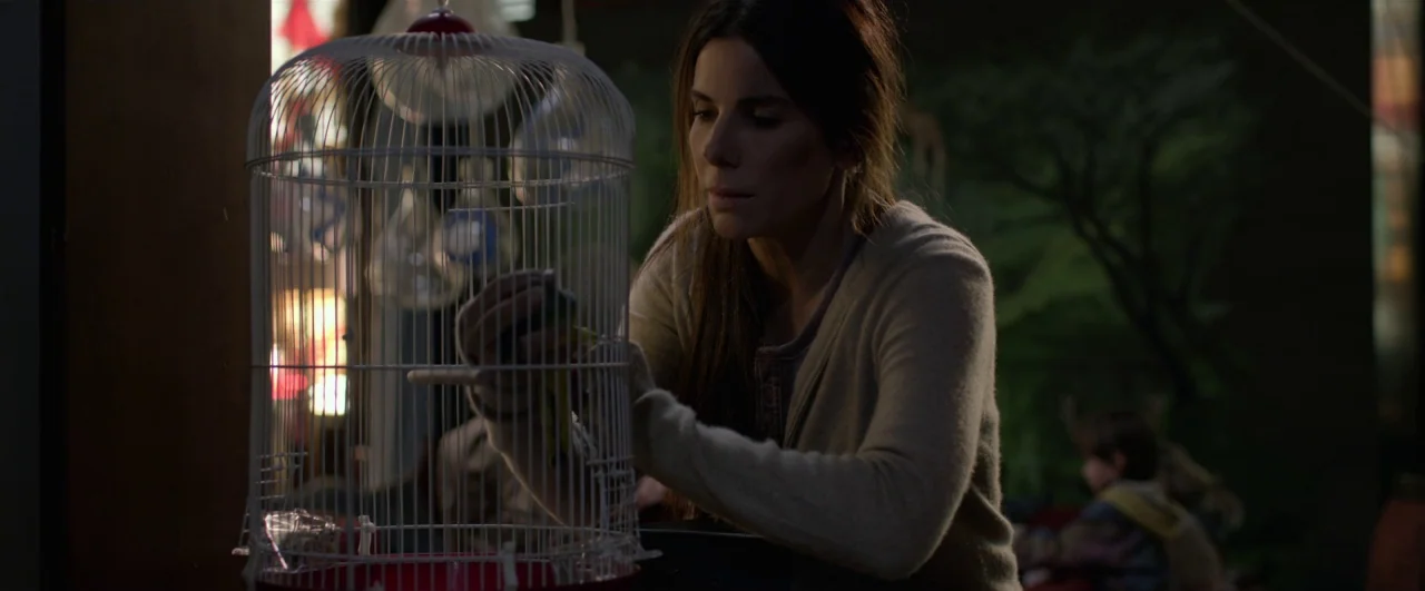 Bird Box (2018) movie still: medium — A dimly lit room provides the backdrop for a person focused on a task. A woman carefully handles a small bird…