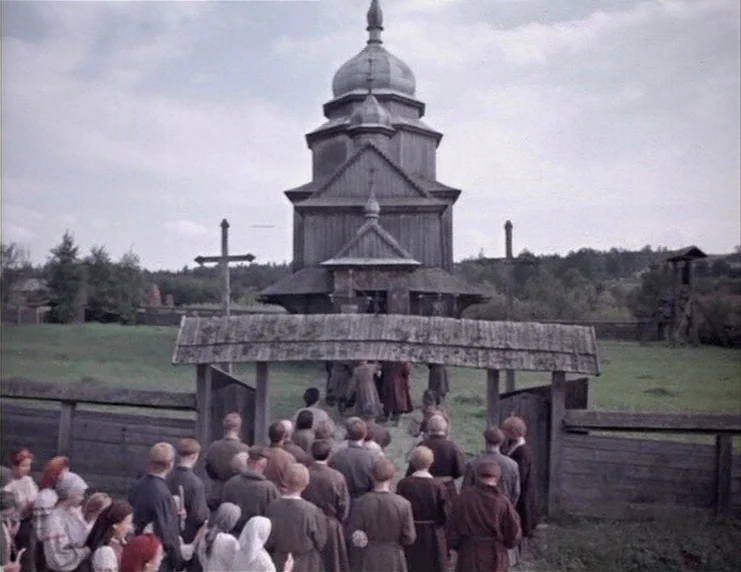 Viy (1967) movie still: wide — A traditional wooden church stands in an open field surrounded by a wooden fence. A group of people dressed in…