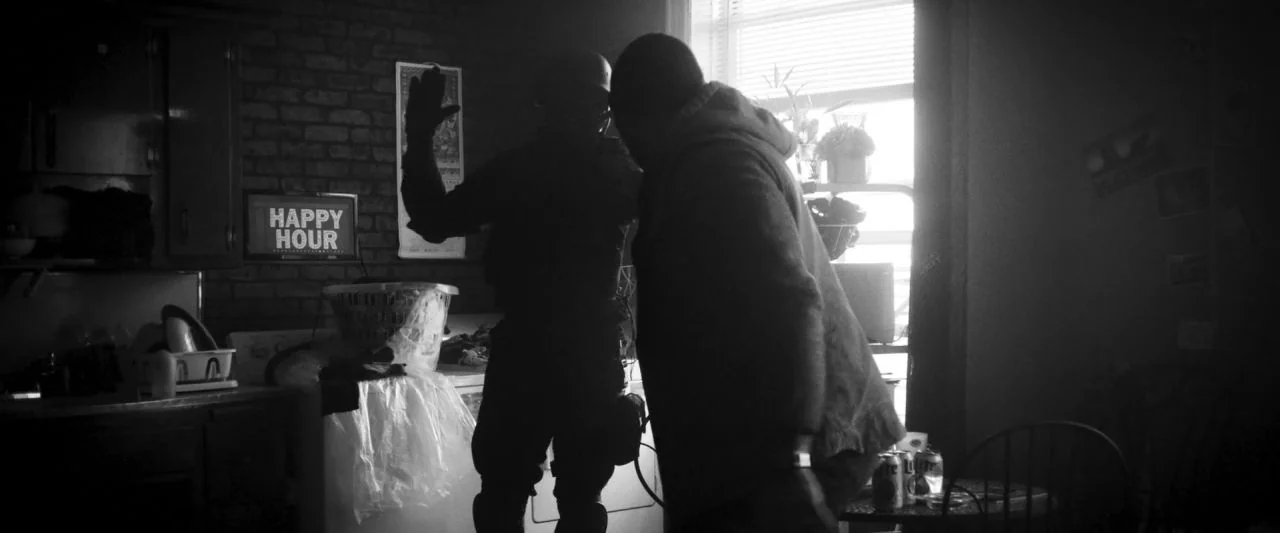 Bushwick (2017) movie still: medium — A dark apartment room with a brick wall and a bright window behind them. Two men face each other with one…