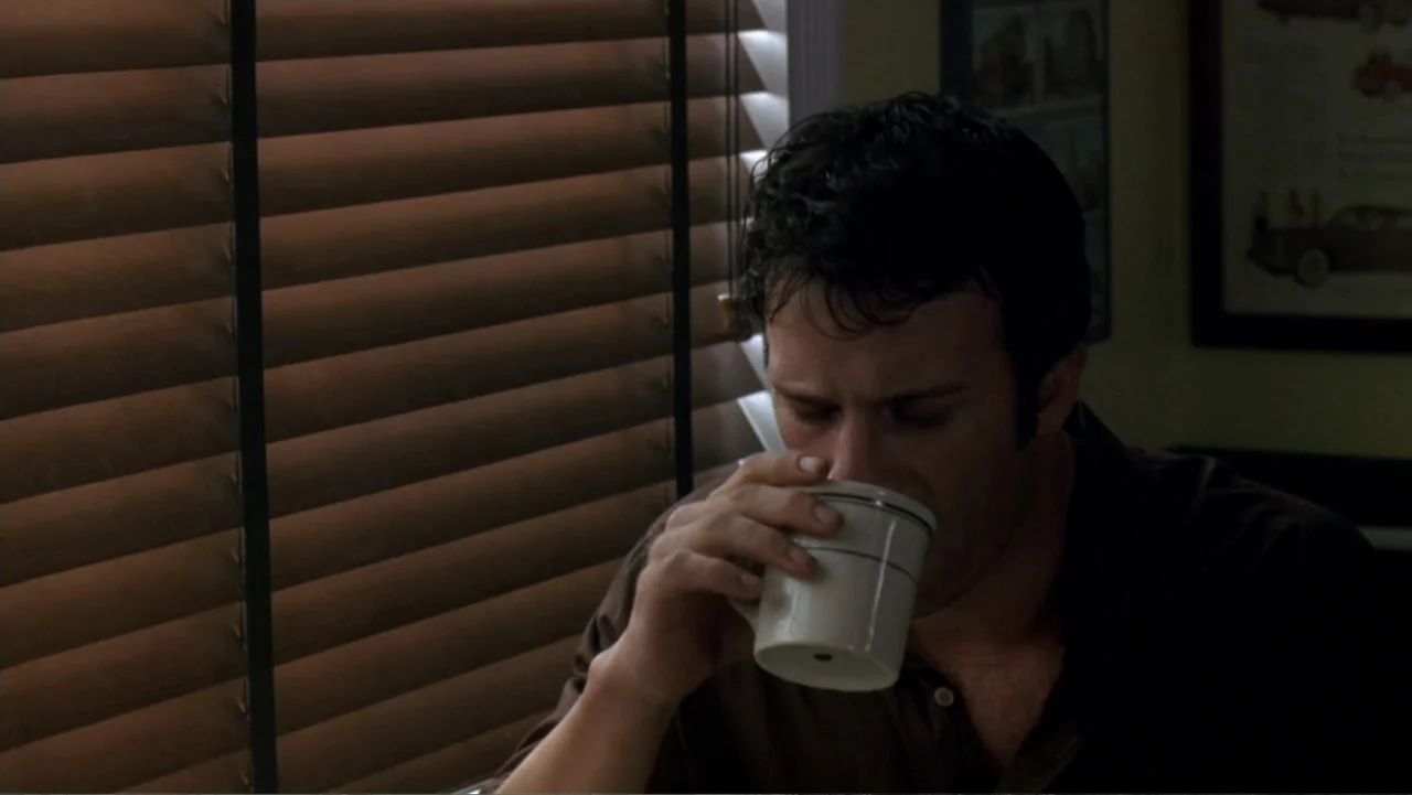 The Punisher (2004) movie still: closeup — A man sits indoors next to a window covered by closed horizontal venetian blinds. He raises a ceramic mug…