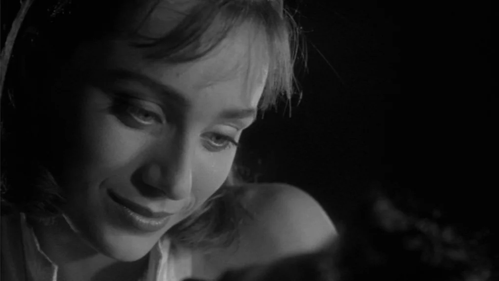 Under The Cherry Moon (1986) movie still: closeup — A dimly lit interior space is swallowed by darkness. A young woman looks down with a faint smile.