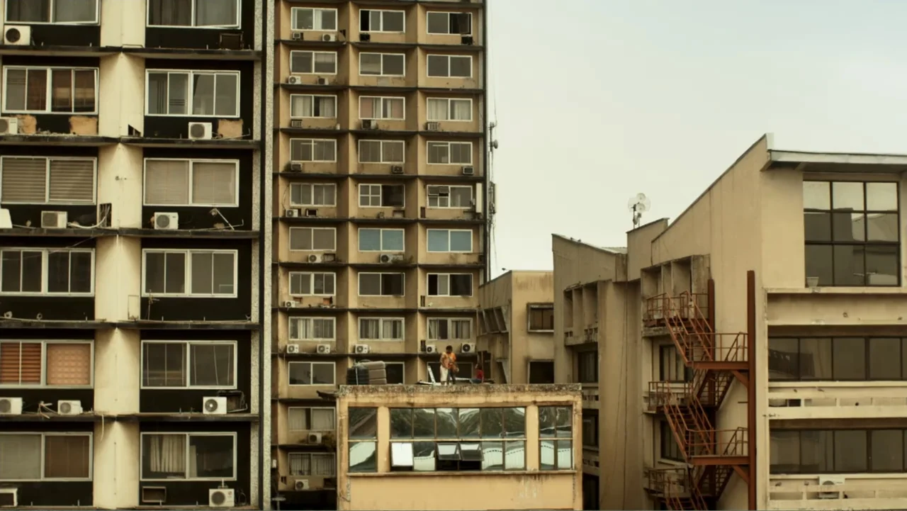 Gangs of Lagos (2023) movie still: wide — An urban apartment complex features beige facades with rows of windows and air conditioners. Two small…