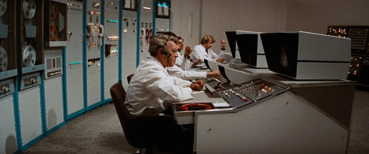 Westworld (1973) movie still: medium — A 1970s technical control room with rows of equipment racks and computer consoles. A group of men in white…