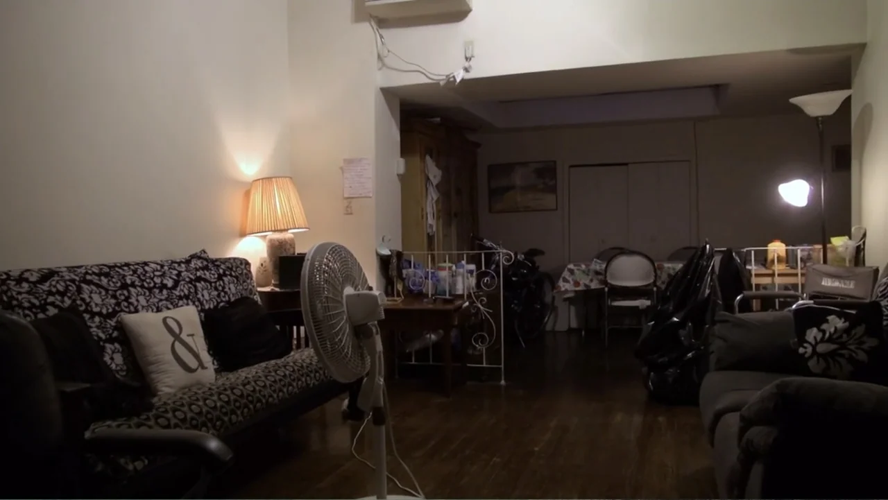 Covid Diaries NYC (2021) movie still: wide — A cluttered residential living room features a patterned sofa and wooden floors at night. Various…