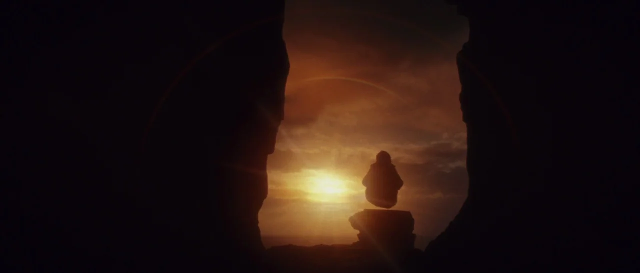 Star Wars: The Last Jedi (2017) movie still: wide — A dark rocky cave opening frames a distant sunset over an horizon line. A lone hooded figure…