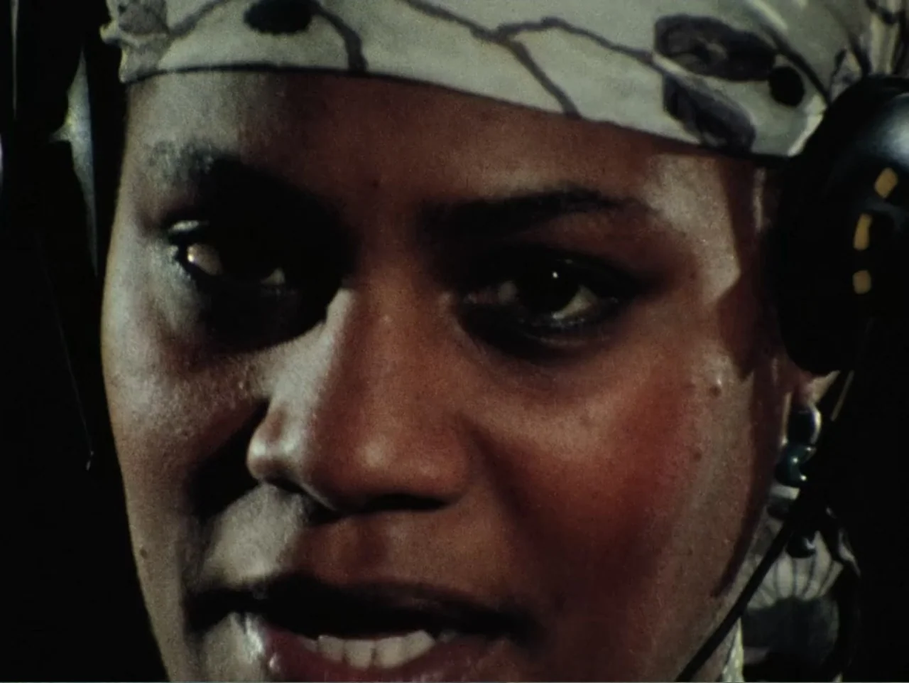 Born in Flames (1983) movie still: closeup — A dimly lit room provides a dark, high-contrast backdrop for a close-up portrait. A woman wearing a…